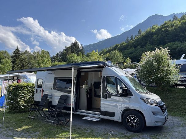 Wohnmobil auf einem Campingplatz, umgeben von Bäumen und Bergen.